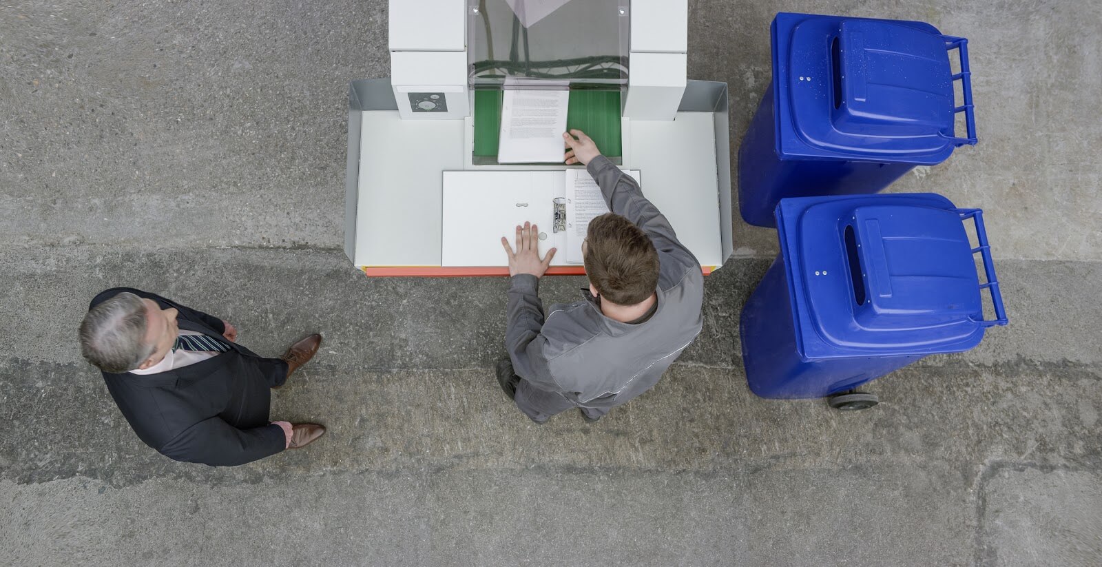 two men looking at documents on a table while two shred bins sit next to it