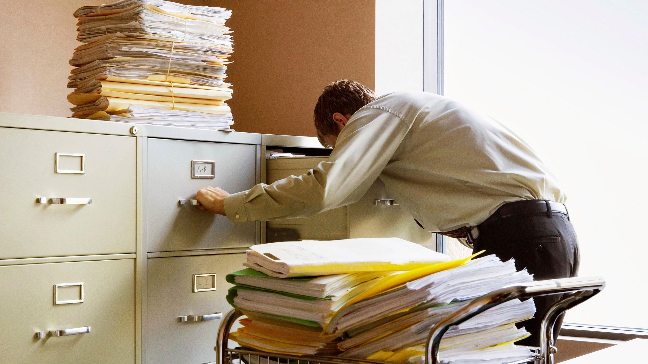 A person in a cluttered office leans into an open filing cabinet surrounded by towering stacks of files and paperwork, conveying a sense of busyness and disorganization.