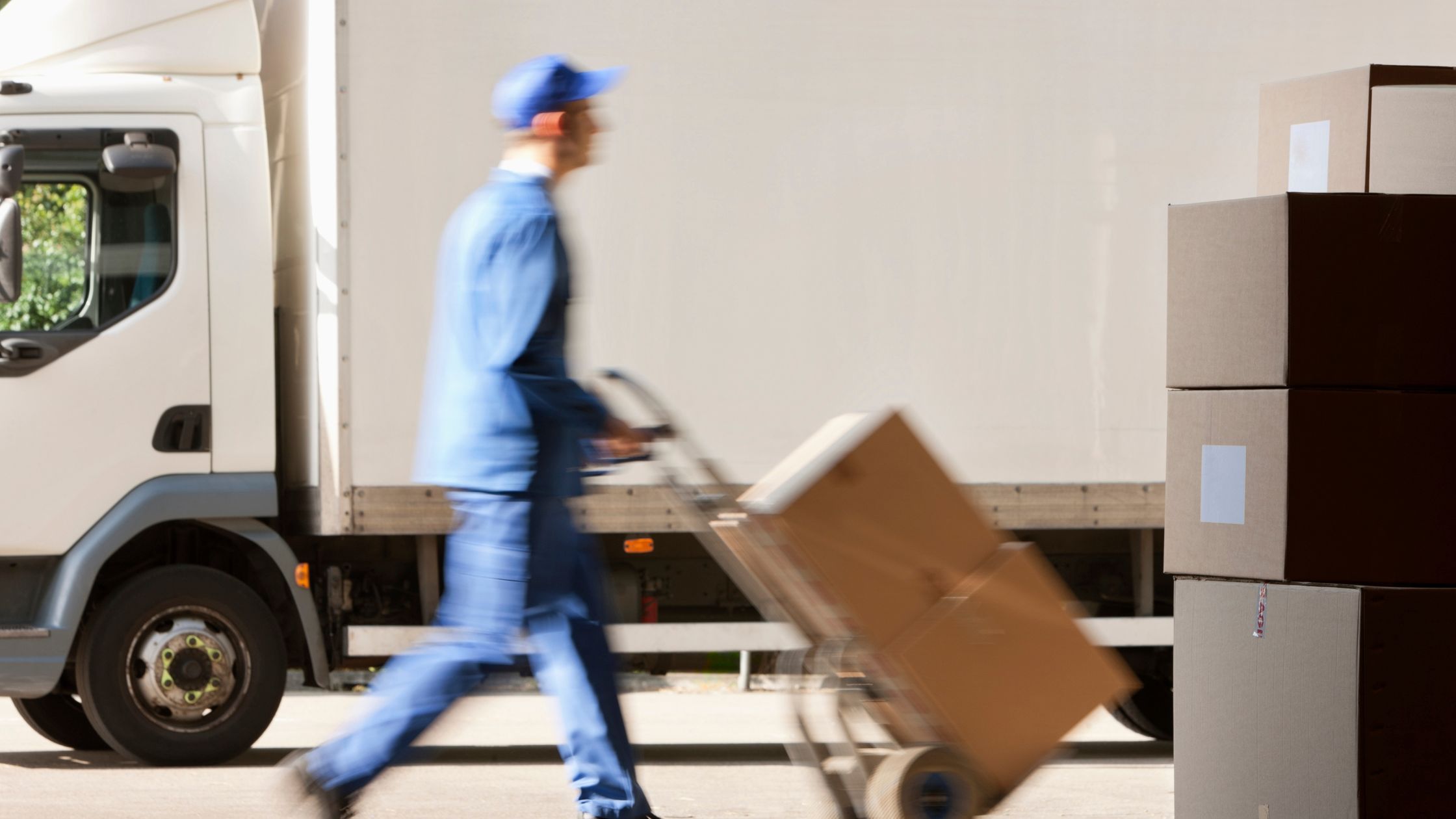 A delivery worker in a blue uniform pushes a hand truck with boxes past a parked white truck. More boxes are stacked nearby, suggesting busy activity.