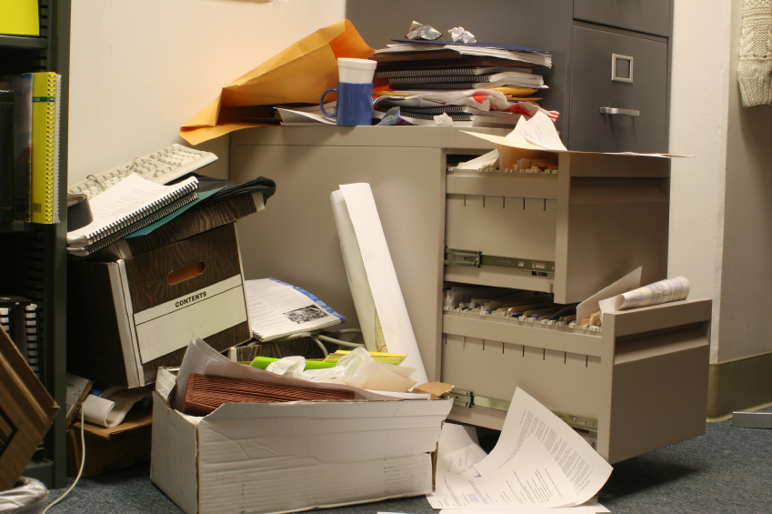 Cluttered office with overflowing filing cabinets, document boxes, and stacks of paper records awaiting organization.