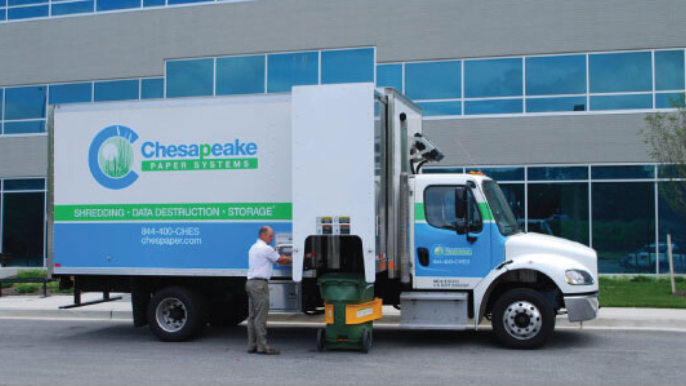 Chesapeake Paper Systems mobile shredding truck providing on-site document destruction outside a commercial building, with an employee securely loading confidential documents for shredding.